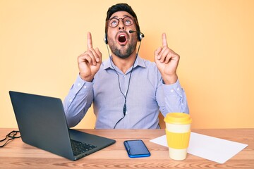 Young hispanic man sitting at the desk wearing operator headset at the call center office amazed and surprised looking up and pointing with fingers and raised arms.