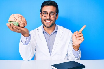 Young hispanic man sitting at the table wearing doctor coat holding brain smiling happy pointing...