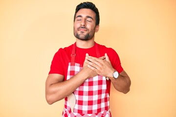Young hispanic man wearing professional baker apron smiling with hands on chest, eyes closed with grateful gesture on face. health concept.