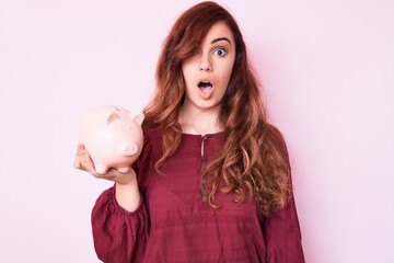 Young beautiful woman holding piggy bank scared and amazed with open mouth for surprise, disbelief face