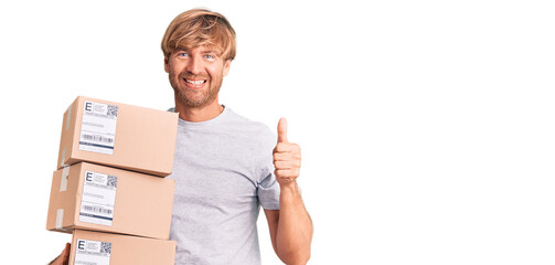 Handsome caucasian man with beard holding delivery packages smiling happy and positive, thumb up doing excellent and approval sign