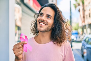 Young hispanic man smiling happy holding breast cancer pink ribbon at city.