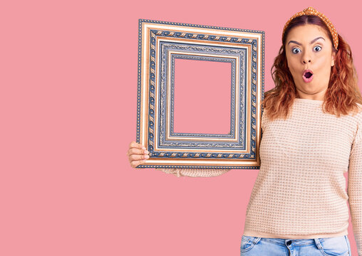 Young latin woman holding empty frame scared and amazed with open mouth for surprise, disbelief face