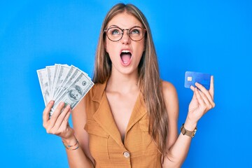 Young blonde girl holding credit car and dollars angry and mad screaming frustrated and furious, shouting with anger looking up.