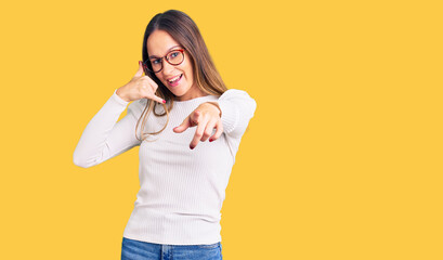 Beautiful brunette young woman wearing casual white sweater and glasses smiling doing talking on the telephone gesture and pointing to you. call me.