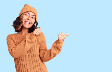 Young beautiful mixed race woman wearing wool sweater and winter hat pointing to the back behind with hand and thumbs up, smiling confident