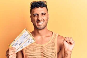 Young hispanic man holding boarding pass screaming proud, celebrating victory and success very excited with raised arm