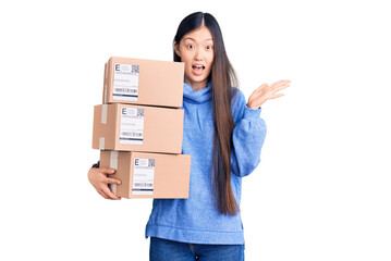 Young beautiful chinese woman holding delivery cardboard boxes celebrating victory with happy smile and winner expression with raised hands