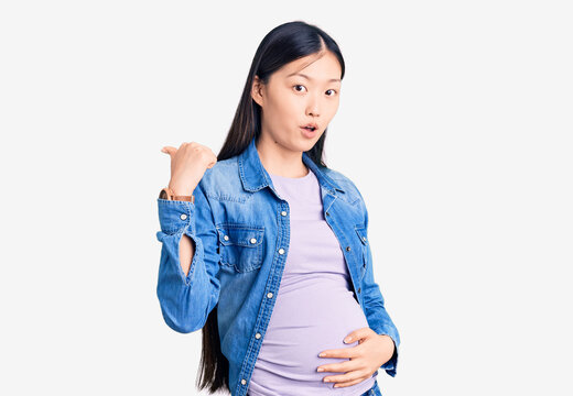 Young beautiful chinese woman pregnant expecting baby surprised pointing with hand finger to the side, open mouth amazed expression.