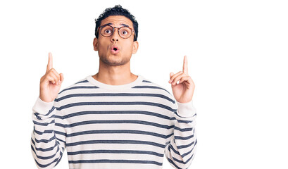 Hispanic handsome young man wearing casual clothes and glasses amazed and surprised looking up and pointing with fingers and raised arms.