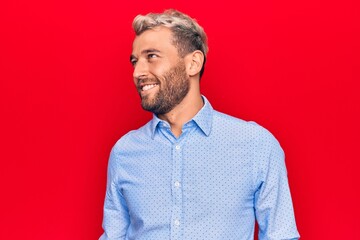 Young handsome blond man wearing casual shirt standing over isolated red background looking to side, relax profile pose with natural face and confident smile.