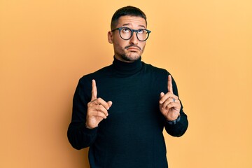 Handsome man with tattoos wearing turtleneck sweater and glasses pointing up looking sad and upset, indicating direction with fingers, unhappy and depressed.