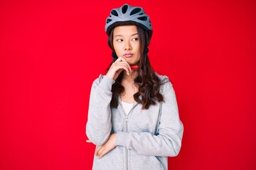 Young beautiful chinese girl wearing bike helmet serious face thinking about question with hand on chin, thoughtful about confusing idea