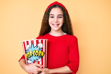 Cute hispanic child girl holding popcorn looking positive and happy standing and smiling with a confident smile showing teeth