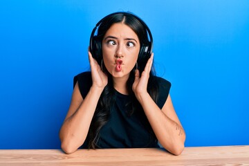 Beautiful brunette young woman listening to music using headphones making fish face with mouth and squinting eyes, crazy and comical.