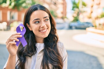 Young hispanic woman smiling happy holding purple ribbon walking at the city.