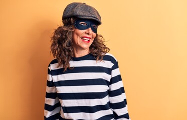 Middle age brunette burglar woman wearing mask and cap standing over yellow background looking to side, relax profile pose with natural face and confident smile.