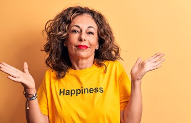 Middle age beautiful woman wearing t-shirt with happiness word over yellow background clueless and confused with open arms, no idea and doubtful face.