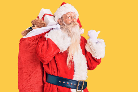 Old senior man with grey hair and long beard wearing santa claus costume holding bag with presents pointing thumb up to the side smiling happy with open mouth