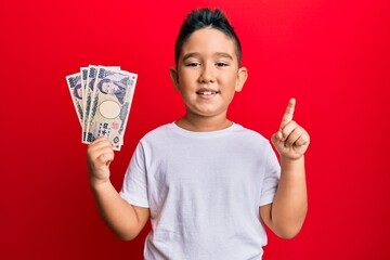 Little boy hispanic kid holding 5000 japanese yen banknotes smiling with an idea or question pointing finger with happy face, number one