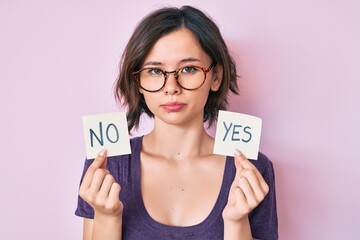 Young beautiful girl holding yes and no reminder relaxed with serious expression on face. simple...