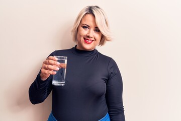 Young beautiful blonde plus size woman drinking glass of water over isolated white background looking positive and happy standing and smiling with a confident smile showing teeth