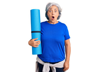 Senior woman with gray hair holding yoga mat scared and amazed with open mouth for surprise, disbelief face