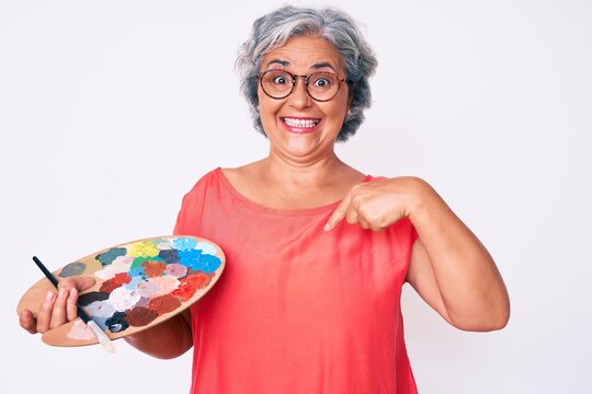 Senior hispanic grey- haired woman holding paintbrush and palette pointing finger to one self smiling happy and proud