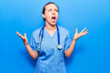 Young blonde woman wearing doctor uniform and stethoscope crazy and mad shouting and yelling with aggressive expression and arms raised. frustration concept.