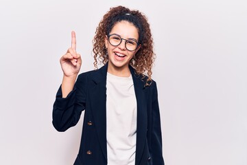 Beautiful kid girl with curly hair wearing business clothes and glasses pointing finger up with successful idea. exited and happy. number one.