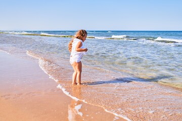 Adorable blonde child on back view wearing summer dress playing on the sand at the beach