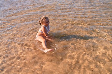 Fototapeta premium Adorable blonde child wearing bikini smiling happy. Sitting on the sand playing with water at the sea