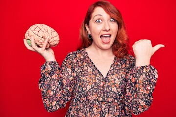Young beautiful redhead woman asking for care memory holding brain over red backgroud pointing...