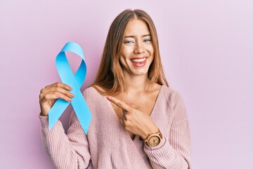 Beautiful young blonde woman holding blue ribbon smiling happy pointing with hand and finger