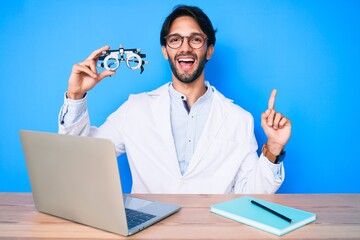 Handsome hispanic optician man at the clinic holding optometry glasses surprised with an idea or question pointing finger with happy face, number one