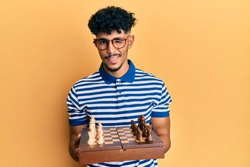 Young arab handsome man holding chess board wearing glasses winking looking at the camera with sexy...