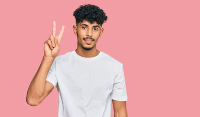 Young arab man wearing casual white t shirt smiling looking to the camera showing fingers doing victory sign. number two.