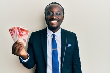 Handsome young black man wearing business suit holding 20 shekels banknotes looking positive and happy standing and smiling with a confident smile showing teeth