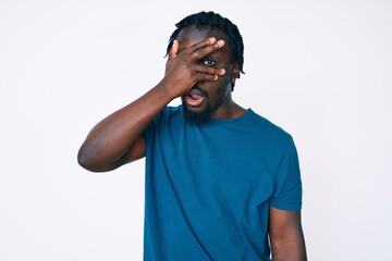 Young african american man with braids wearing casual clothes peeking in shock covering face and eyes with hand, looking through fingers with embarrassed expression.