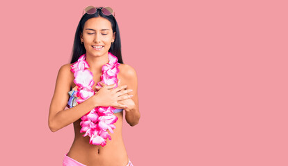 Young beautiful latin girl wearing bikini and hawaiian lei smiling with hands on chest with closed eyes and grateful gesture on face. health concept.