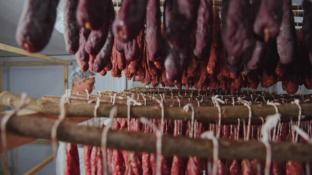At meat factory worker inspects salami and sausage smoking process in smockhouse. Meat sausages production. Farming business. Quality control.