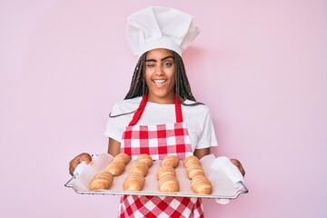 Young african american woman with braids wearing baker uniform holding homemade bread winking...