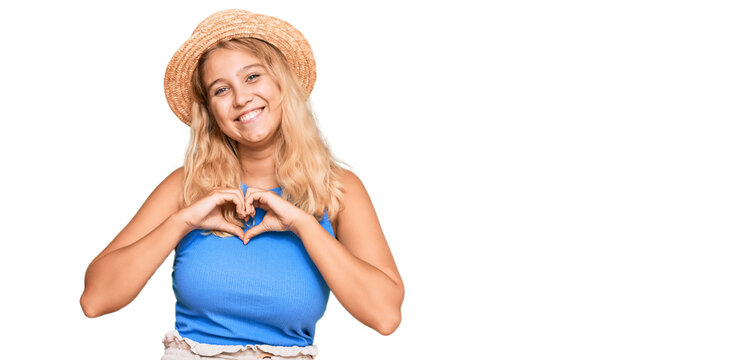 Young blonde girl wearing summer hat smiling in love showing heart symbol and shape with hands. romantic concept.