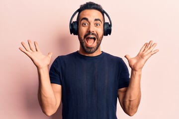 Young hispanic man listening to music using headphones celebrating victory with happy smile and winner expression with raised hands