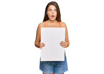 Young caucasian woman holding blank empty banner scared and amazed with open mouth for surprise, disbelief face