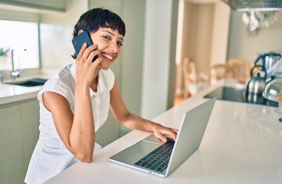 Beautiful brunette woman with short hair at home using computer laptop speaking on the phone