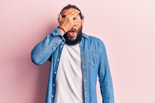 Young arab man wearing casual clothes peeking in shock covering face and eyes with hand, looking through fingers afraid