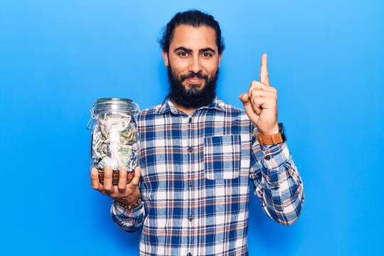 Young arab man holding jar with savings smiling with an idea or question pointing finger with happy face, number one