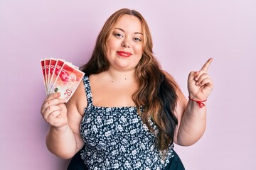 Plus size caucasian young woman holding 20 israel shekels banknotes smiling happy pointing with...
