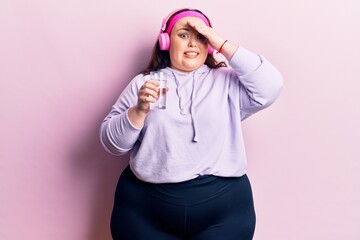 Obraz na płótnie Canvas Young plus size woman drinking glass of water using headphones stressed and frustrated with hand on head, surprised and angry face